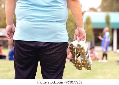 Fat Man Holding Football Boots And Stands Watching Soccer Practice.