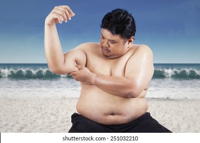 Fat Man Holding His Flabby Biceps, Shot Outdoors At Beach