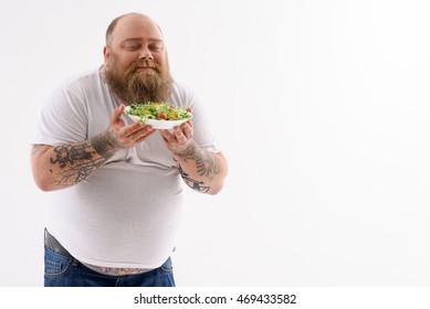 Fat man is holding plate of salad and smelling it with pleasure. He is standing with closed eyes. Isolated and copy space in left side