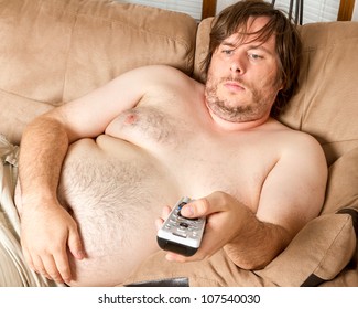 Fat man is laying on the couch topless watching the TV. Guy is overweight and quite lazy looking.