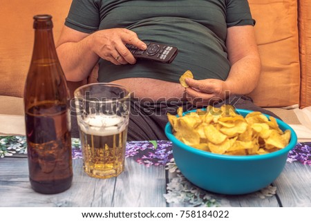 A fat man suffers from overweight and eats chips, drinks alcohol and looks in the telly. Concept: Healthcare