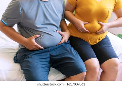 Fat man and woman holding their big belly waistline sitting on the bed suffering from extra weight. Couple heavy body size worry problem bad healthy in the bedroom.Overweight loss unhealthy concept.