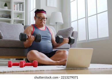 Fat middle-aged asian man in sportswear using dumbbell exercising following trainer online via computer laptop at home