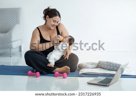 Fat mom in sportswear pausing online fitness class on laptop to bottle feed baby, doing exercise on floor mat at home