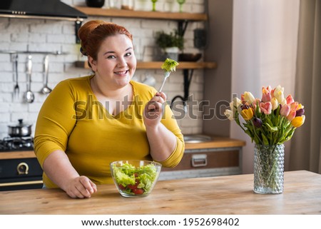 Fat obese plump young caucasian woman eating salad for healthy diet and eating habits in the kitchen. Chubby plus size woman having salad for breakfast