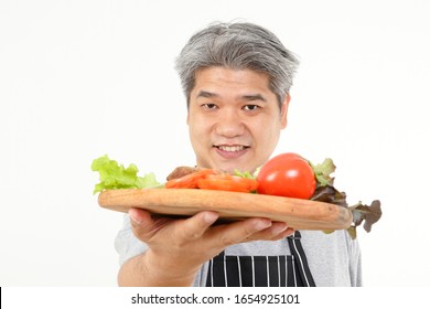 Fat Old Man Holds A Wooden Tray With Fresh Vegetables For Water To Cook. The Concept Of Healthy Eating For The Elderly. White Background