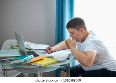 fat overweight boy eat junk food while doing homework, young caucasian boy sit at desk with notebooks, laptop and books. at home