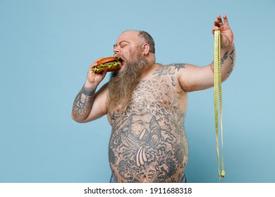 Fat pudge obese chubby overweight man has tattooed naked bare big belly eating hold fast food burger measure tape isolated on blue color background studio. Weight loss obesity unhealthy diet concept