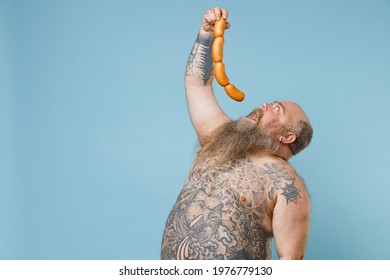 Fat pudge obese chubby overweight man has tattooed naked bare big belly hold in hand eating sausages isolated on blue color background studio portrait. Weight loss obesity unhealthy diet concept