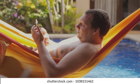 Fat Sad Man With Bare Chest Lies In Hammock Uses Mobile Phone By Swimming Pool.