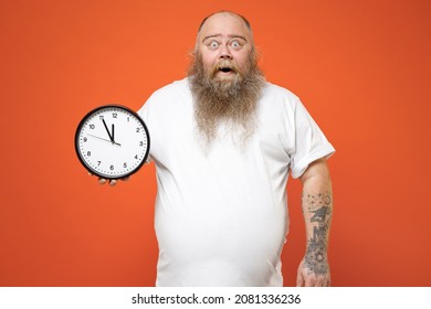 Fat shocked pudge obese chubby overweight tattooed blue-eyed bearded man 30s has big belly wear white t-shirt hold in hands clock look camera isolated on red orange color background studio portrait.