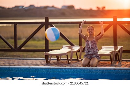 Fat Tanned Happy Girl With Dark Hair Braided In Bun In Bright Summer Suit Is Playing Near Pool With Inflatable Ball, Against The Background Of Golden Summer Evening Sun