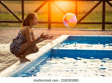 Fat Tanned Happy Girl With Dark Hair Braided In Bun In Bright Summer Suit Is Playing Near Pool With Inflatable Ball, Against The Background Of Golden Summer Evening Sun