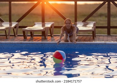 Fat Tanned Happy Girl With Dark Hair Braided In Bun In Bright Summer Suit Is Playing Near Pool With Inflatable Ball, Against The Background Of Golden Summer Evening Sun