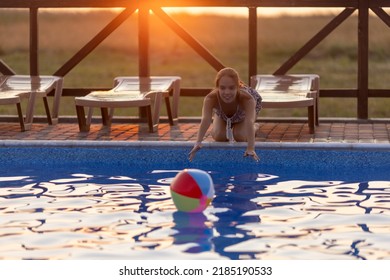 Fat Tanned Happy Girl With Dark Hair Braided In Bun In Bright Summer Suit Is Playing Near Pool With Inflatable Ball, Against The Background Of Golden Summer Evening Sun