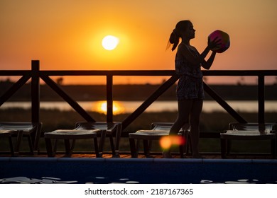 Fat Tanned Happy Girl With Dark Hair Braided In Bun In Bright Summer Suit Is Playing Near Pool With Inflatable Ball, Against The Background Of Golden Summer Evening Sun