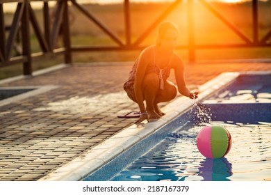 Fat Tanned Happy Girl With Dark Hair Braided In Bun In Bright Summer Suit Is Playing Near Pool With Inflatable Ball, Against The Background Of Golden Summer Evening Sun