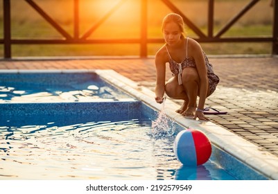 Fat Tanned Happy Girl With Dark Hair Braided In Bun In Bright Summer Suit Is Playing Near Pool With Inflatable Ball, Against The Background Of Golden Summer Evening Sun