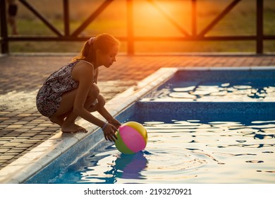 Fat Tanned Happy Girl With Dark Hair Braided In Bun In Bright Summer Suit Is Playing Near Pool With Inflatable Ball, Against The Background Of Golden Summer Evening Sun