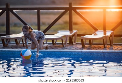 Fat Tanned Happy Girl With Dark Hair Braided In Bun In Bright Summer Suit Is Playing Near Pool With Inflatable Ball, Against The Background Of Golden Summer Evening Sun