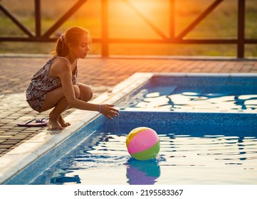 Fat Tanned Happy Girl With Dark Hair Braided In Bun In Bright Summer Suit Is Playing Near Pool With Inflatable Ball, Against The Background Of Golden Summer Evening Sun