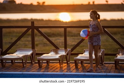 Fat Tanned Happy Girl With Dark Hair Braided In Bun In Bright Summer Suit Is Playing Near Pool With Inflatable Ball, Against The Background Of Golden Summer Evening Sun