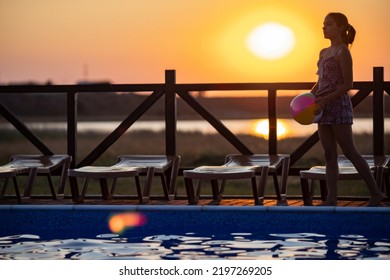 Fat Tanned Happy Girl With Dark Hair Braided In Bun In Bright Summer Suit Is Playing Near Pool With Inflatable Ball, Against The Background Of Golden Summer Evening Sun