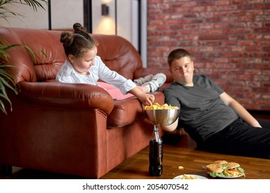 Fat Teenager Boy Sharing Crisps With Sister Sitting On Floor, Having Rest Together, Kid Girl Spend Leisure Time With Brother At Home, In Living Room, Watching Tv. Unhealthy Lifestyle