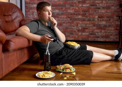 Fat teenager boy sitting on floor in living room, side view. Overweight obese caucasian child in casual clothes enjoy leading unhealthy lifestyle, eat junk food and play video games. - Shutterstock ID 2090350039