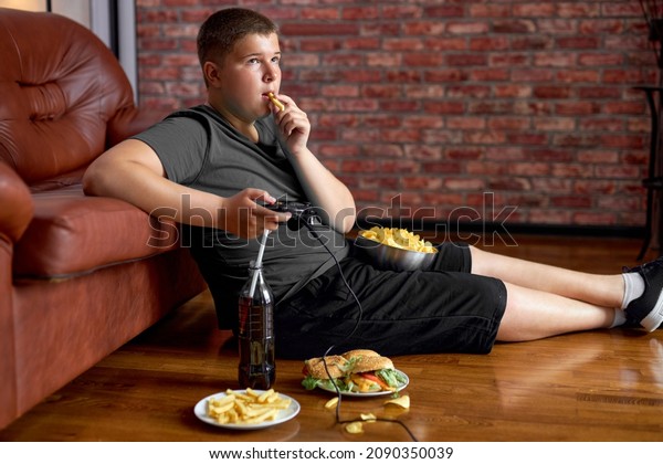 Fat\
teenager boy sitting on floor in living room, side view. Overweight\
obese caucasian child in casual clothes enjoy leading unhealthy\
lifestyle, eat junk food and play video\
games.