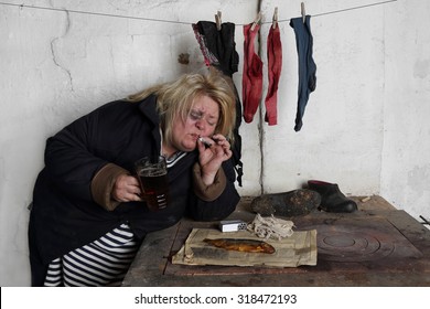 Fat terrible woman with bruise on face smokes cigarette leaning on the stove with mug of beer and dried fish on old yellow newspaper under hung out to dry laundry - alcoholism and scum concept