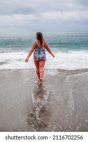 A fat woman in a bathing suit enters the water during the surf. Alone on the beach, Gray sky in the clouds, swimming in winter.