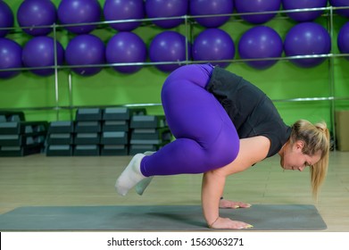 Fat woman is engaged in pilates in a fitness gym. A girl with a lot of overweight trying to lose weight with the help of yoga, stands in a raven pose. The blonde performs a bokasana.