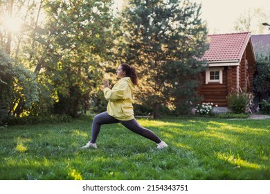 Fat woman of middle age doing yoga exercises in sportswear looking away on fresh grass on backyard with wooden country house and tall trees in background. Body positive.