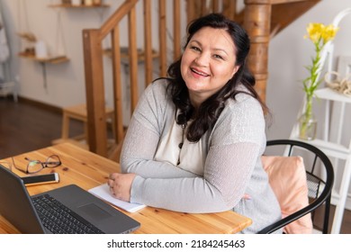 A fat woman is sitting at a table with a laptop at home, in the office. She smiles. - Shutterstock ID 2184245463