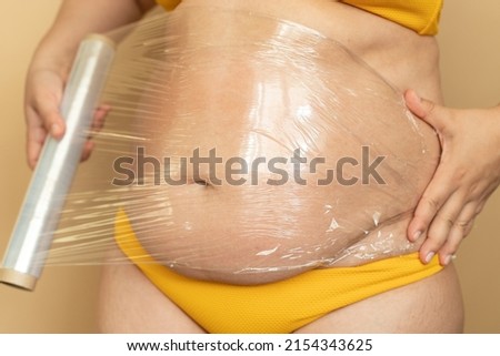 Fat woman in yellow swimsuit wrap thick stomach with cling film closeup, beige background. Slimming, fighting overweight, obesity and cellulite. Plus size people and body positive concept.