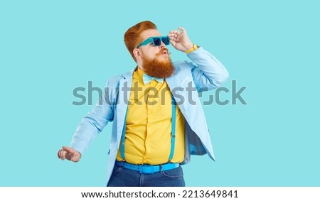 Fat young man in funky modern outfit having fun at disco party. Funny fashion guy with ginger beard and mustache wearing trendy blue specs, jacket, yellow shirt and bowtie dancing on blue background