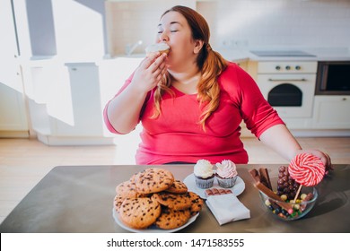 Fat young woman in kitchen sitting and eating sweet food. Gluttony. Fat pancakes and cookies on table. Plus size model in kitchen. Body positive.