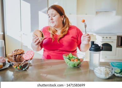 Fat Young Woman In Kitchen Sitting And Eating Junk Food. Holding Burger And Tomato On Fork. Regret And Unhappiness. Body Positive. Sweets On Left. Healthy Food On Right Side.