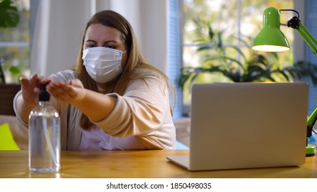 Fat Young Woman Wearing Protective Mask Disinfecting Hands With Antiseptic Working On Laptop At Home. Overweight Lady Cleaning Hands With Sanitizer Sitting At Desk With Computer