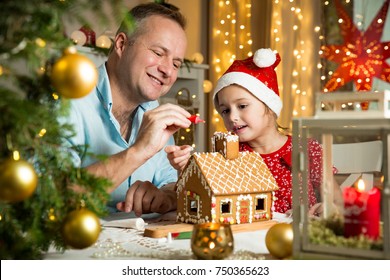 Father and adorable daughter in red hat building gingerbread house together. Beautiful decorated room with lights and Christmas tree, table with candles and lanterns. Happy family celebrating holiday.