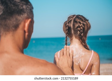 A Father Applies Protective Cream To His Daughter's Back At The Beach. A Man Holds Sunscreen Lotion On A Child's Body. Cute Little Girl With Sunscreen By The Sea. Copy Space.