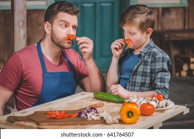 Father In Apron And Cute Little Son Having Fun With Pepper Moustaches, Dad And Son Cooking Concept