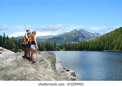 Father With Arms Around His Family Looking At Beautiful Summer Mountains Landscape, On Hiking Trip. People Standing On  Top Of  Mountain Rock. Rocky Mountains National Park, Colorado ,USA.