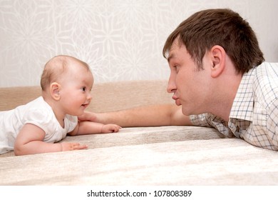 Father And Baby Daughter, Talking, Smiling,  Lying On The Bed And Looking Eye To Eye With Each Other.