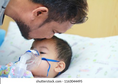 Father With Beard And Mustache Kiss His Little Son While He Had  Nebulizations,Sick Boy Rest On Patien Bed And Has Inhalation Therapy By The Mask Of Inhaler.Sick Or Cancer Child Awareness.