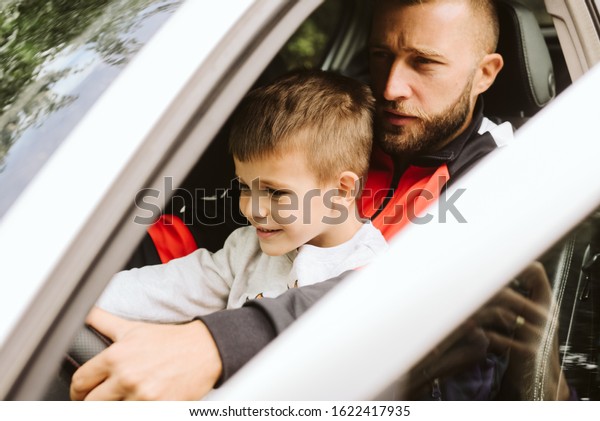 A father with a beard teaches his son to\
drive a car, a son sits in his father\'s\
lap