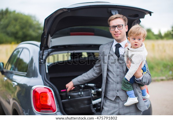 Father businessman dressed in suit tie and\
sunglasses, holding a baby son in her arms and pulls the carriage\
of baggage black car