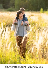 Father Carries His Son On Shoulders. Family Time Outdoors. Man With Child On Autumn Field At Sunset. Dad And Son Explores Nature Together.