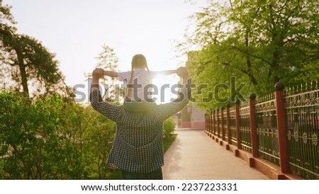 father carries little daughter shoulders park sunset. happy family. dad carries child kid back city park sun. happy family concept. parent small child arms. little superhero rides father shoulders.